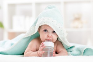 Adorable child baby drinking water from bottle. Little girl wrapped bathing towel lying on bed.